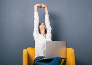 A woman sits on a sofa with laptop on a lap and stretches her arms up in the air